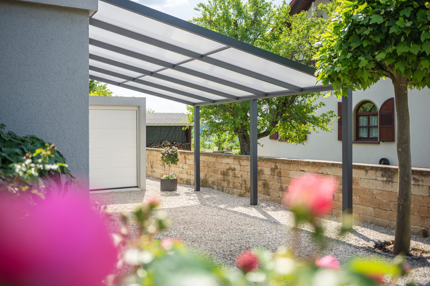 Ein Kies-Stellplatz vor einer Garage, umgeben von Grünflächen und mit einem eleganten anthrazitfarbenen Terrassendach Pro beschattet. Im Hintergrund ist das Nachbarshaus zu sehen.